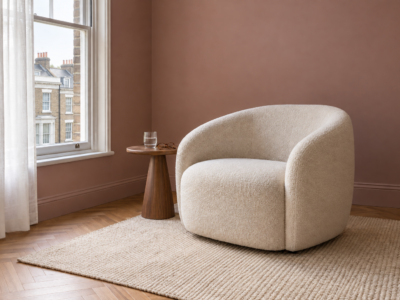 An ivory boucle armchair beside a small walnut table, by a sash window in a living room with dusty rose walls.