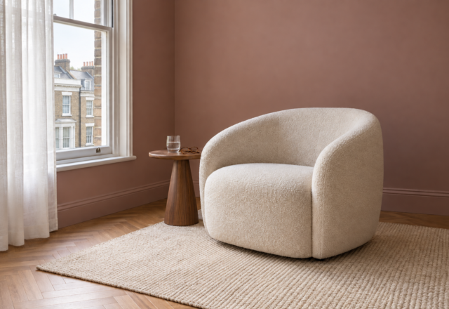 An ivory boucle armchair beside a small walnut table, by a sash window in a living room with dusty rose walls.