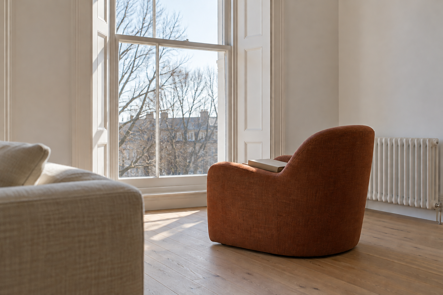 A low rust linen armchair seen from behind, turned to face a tall sash window with its back to the rest of the living room.