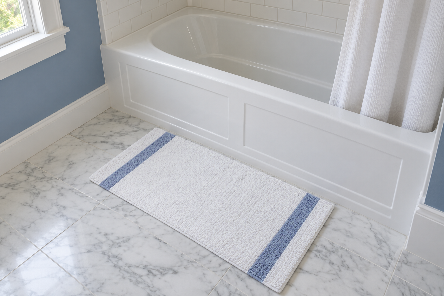 A view from above of a white bath mat lying along the front of a white tub at its middle, shorter than the tub, on a marble floor.