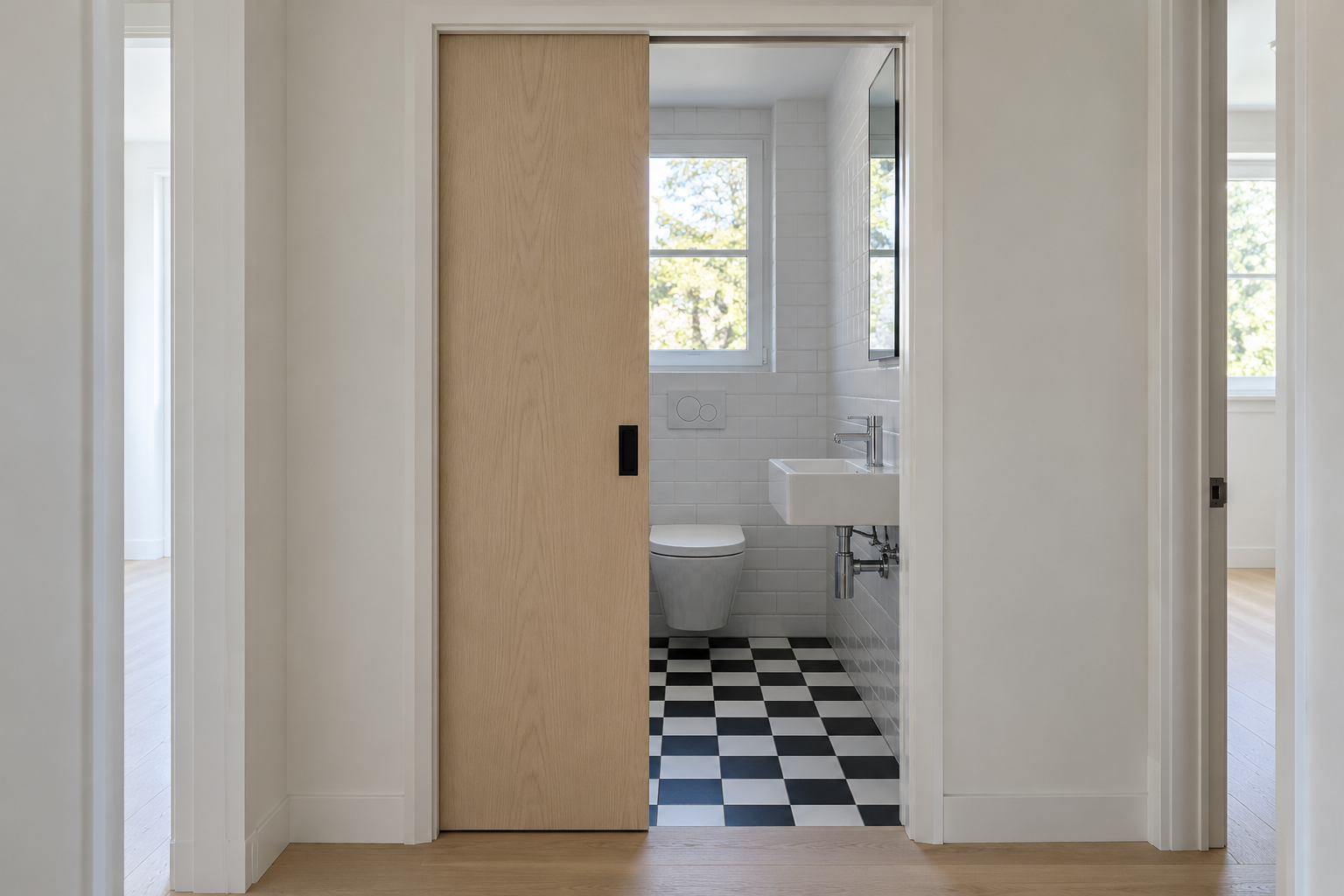 A pale wood pocket door half open, sliding into the wall, with a small bathroom and a checkerboard floor beyond.