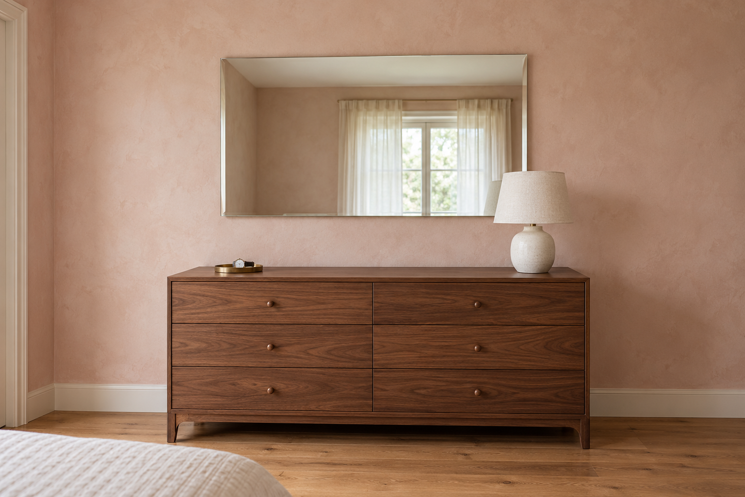 A frameless rectangular mirror with a beveled edge hung a few inches above a walnut dresser on a pale clay plaster wall, reflecting a window.