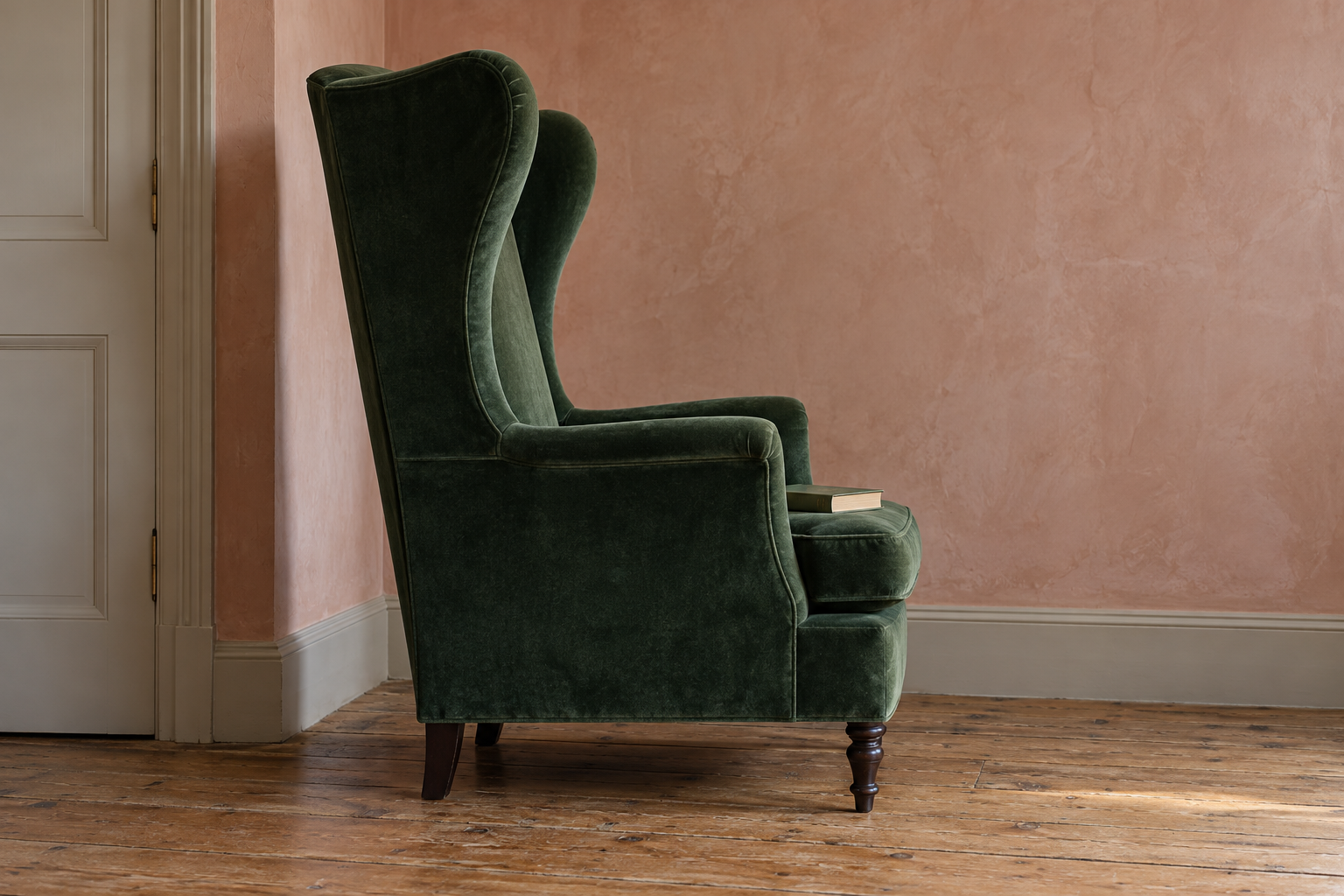 A tall bottle green velvet wing chair seen in profile against a pink plaster wall, its deep wings curving forward.