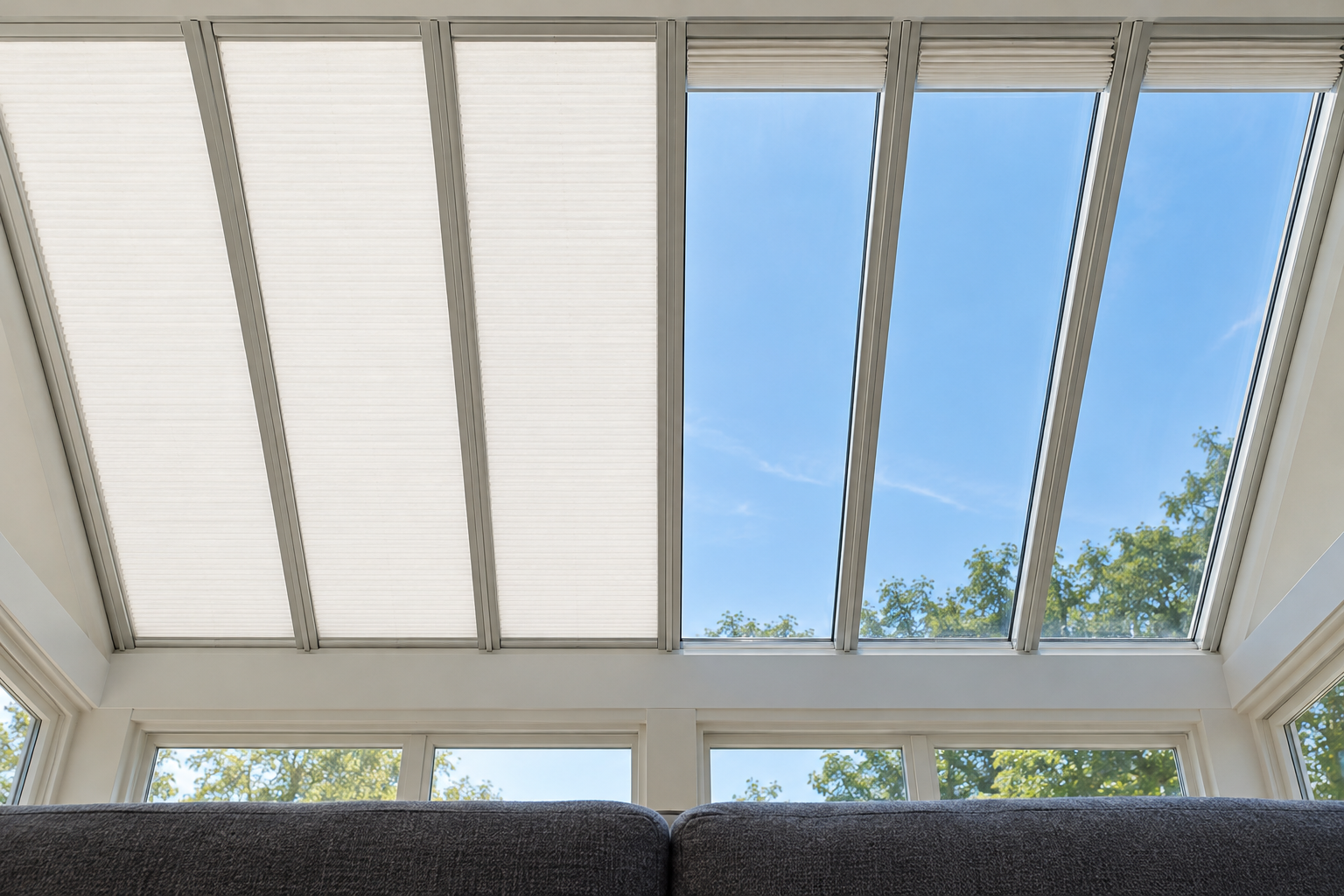 A glass sunroom roof seen from below, with white pleated shades drawn across half of the roof panels in side tracks, and a charcoal sofa at the bottom of the frame.
