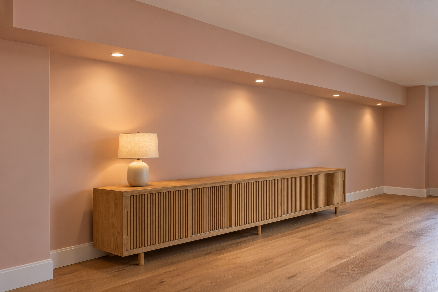 A basement wall with a drywall soffit running its full length near the ceiling, small downlights set in it, and a long low oak cabinet underneath.