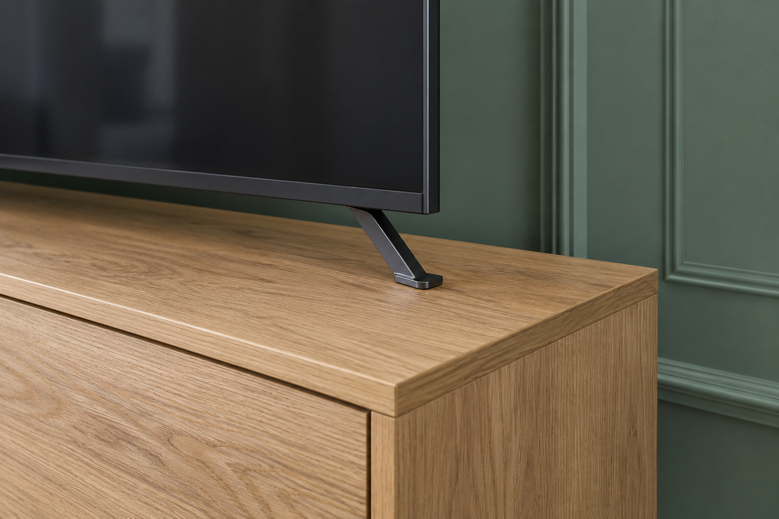 A close view of one end of a media console, showing a television's slim metal foot sitting wholly on the oak top a few inches in from the console's edge.