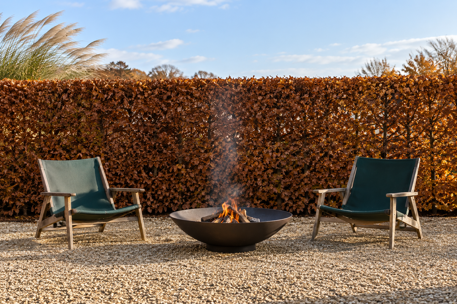 A fire bowl and two chairs on gravel in front of a tall clipped beech hedge with copper leaves.