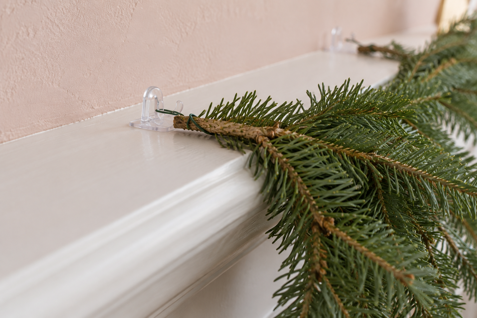 A small clear adhesive hook on top of a white mantel shelf holding a loop of green wire around the stem of a garland.