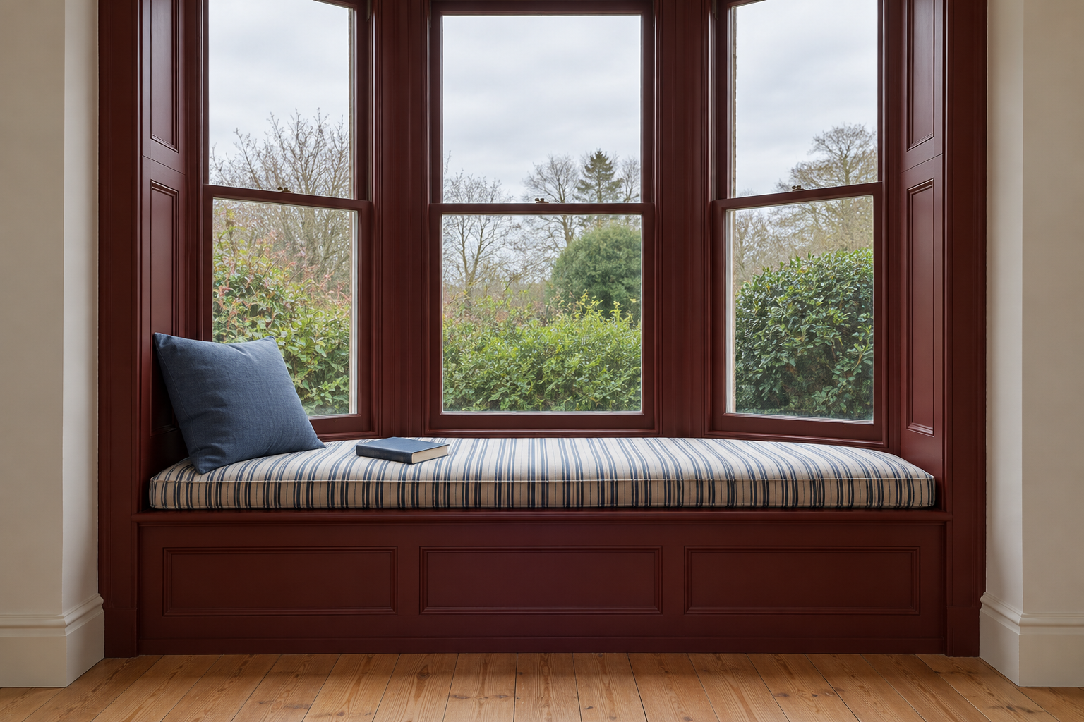 A deep window seat built across a bay window, painted oxblood, with a long striped cushion and a pillow at one end.