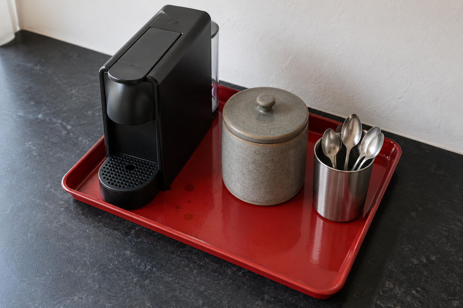 A small coffee machine, a canister, and spoons on a red enamel tray on a black stone counter.