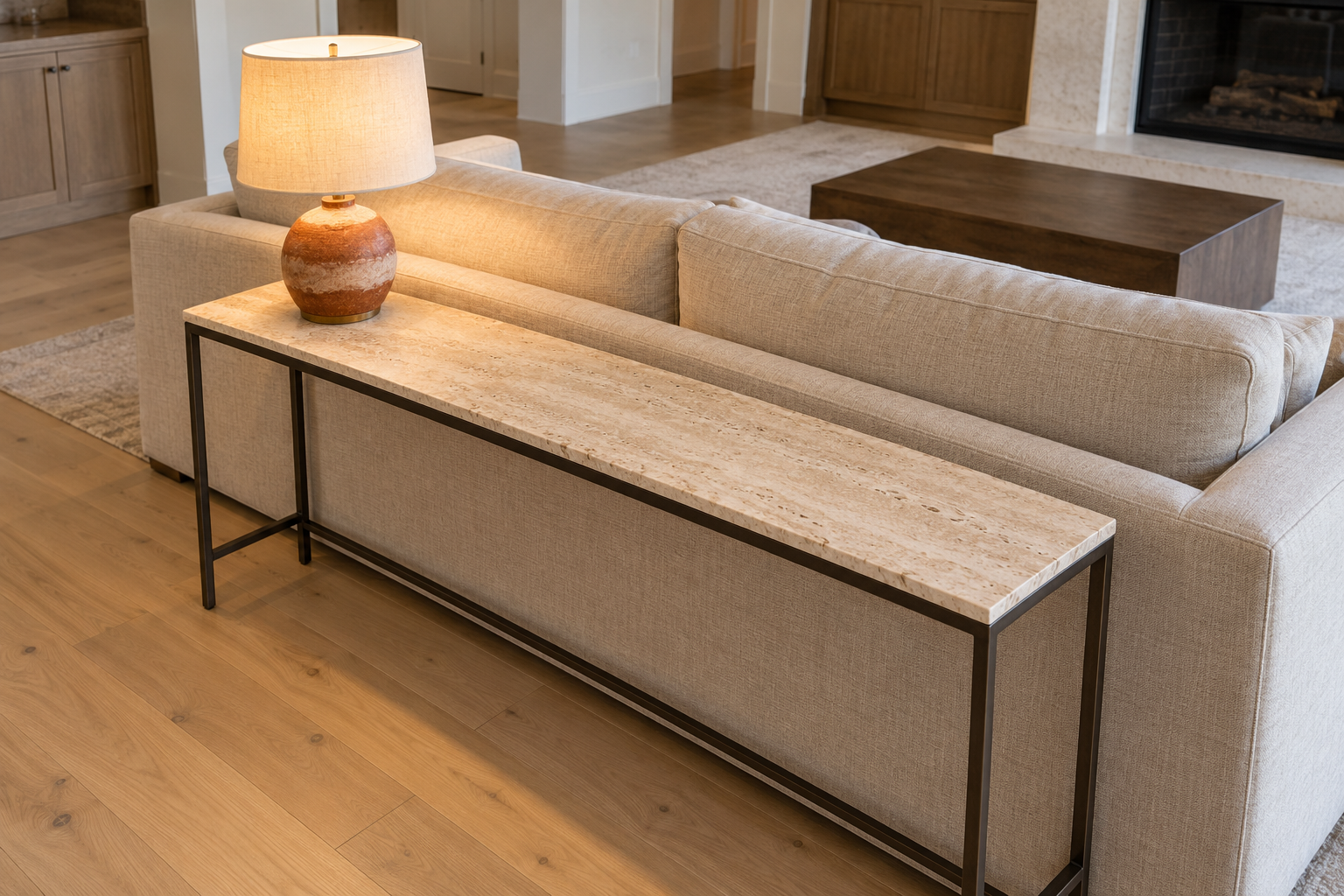 A view from above and behind a sofa table, showing its narrow travertine top ending inside the arms of the linen sofa in front of it.