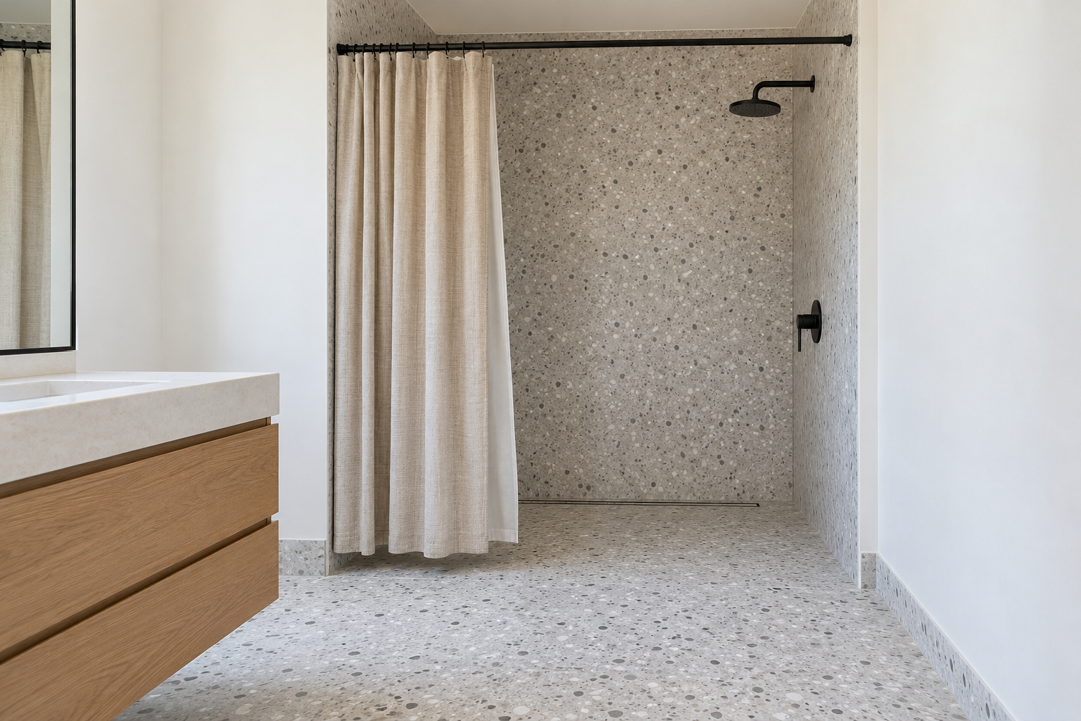 A curbless walk-in shower in pale terrazzo with a natural linen-look curtain hung from a slim rod, its hem about an inch above the floor, and a linear drain along the back.