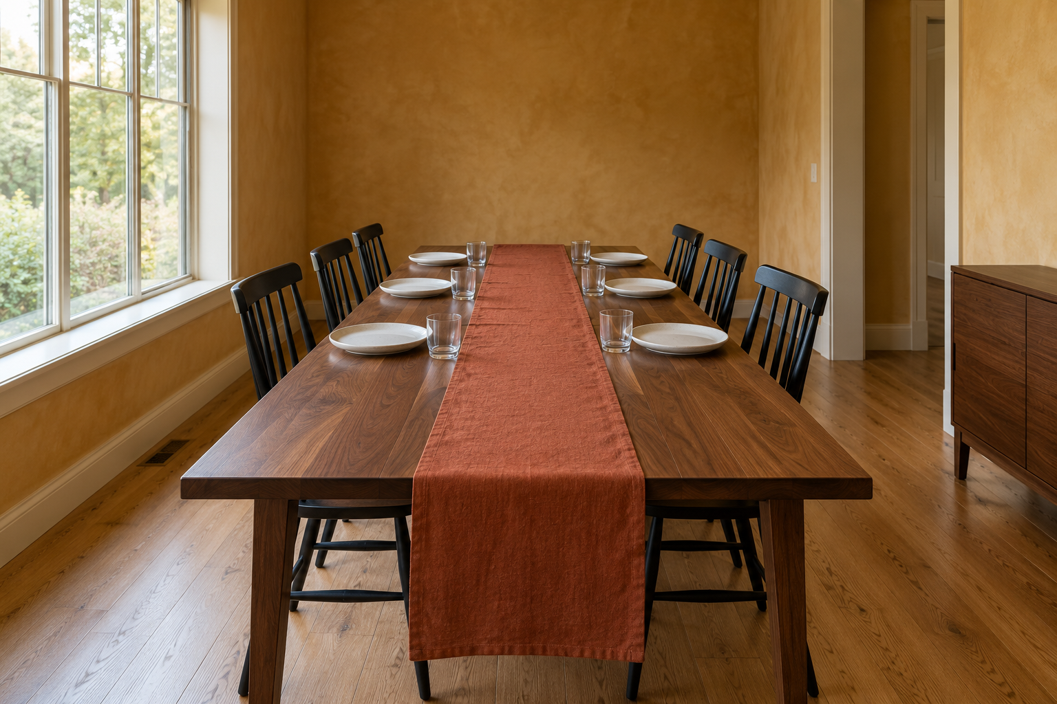 A walnut dining table set with white plates and glasses, with a rust linen runner down the middle that hangs well over the near end.