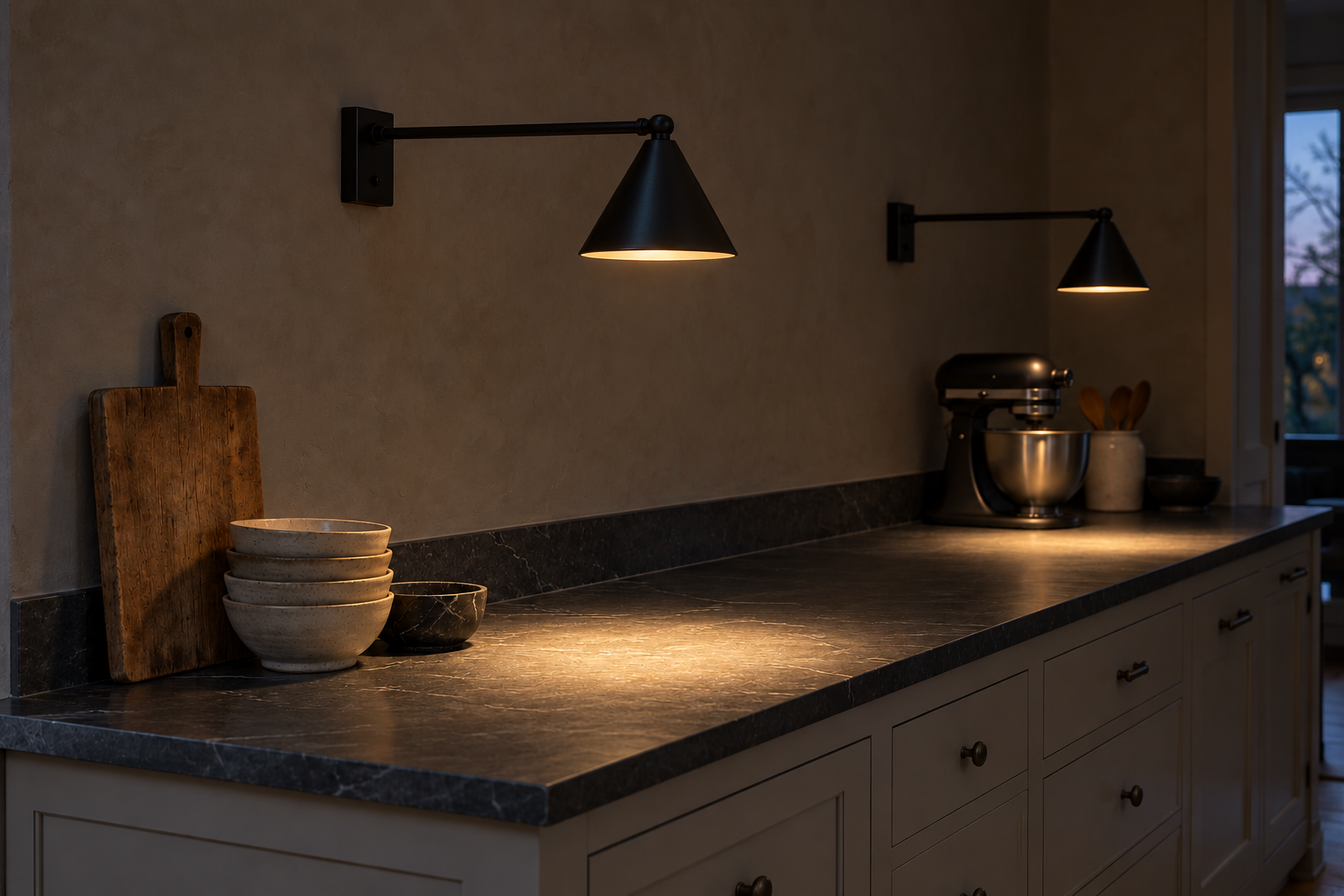 Two blackened steel wall lights on swinging arms reaching over a soapstone kitchen counter, against a warm gray limewashed wall.