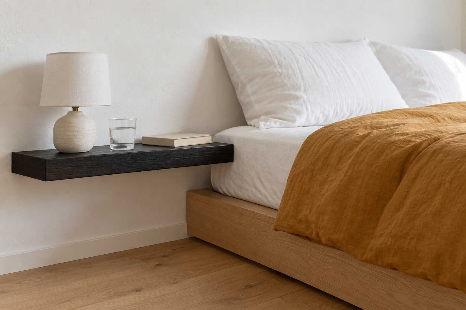 A black oak floating shelf beside a low oak bed, its top level with the top of the mattress, holding a small lamp, a glass of water, and a book.