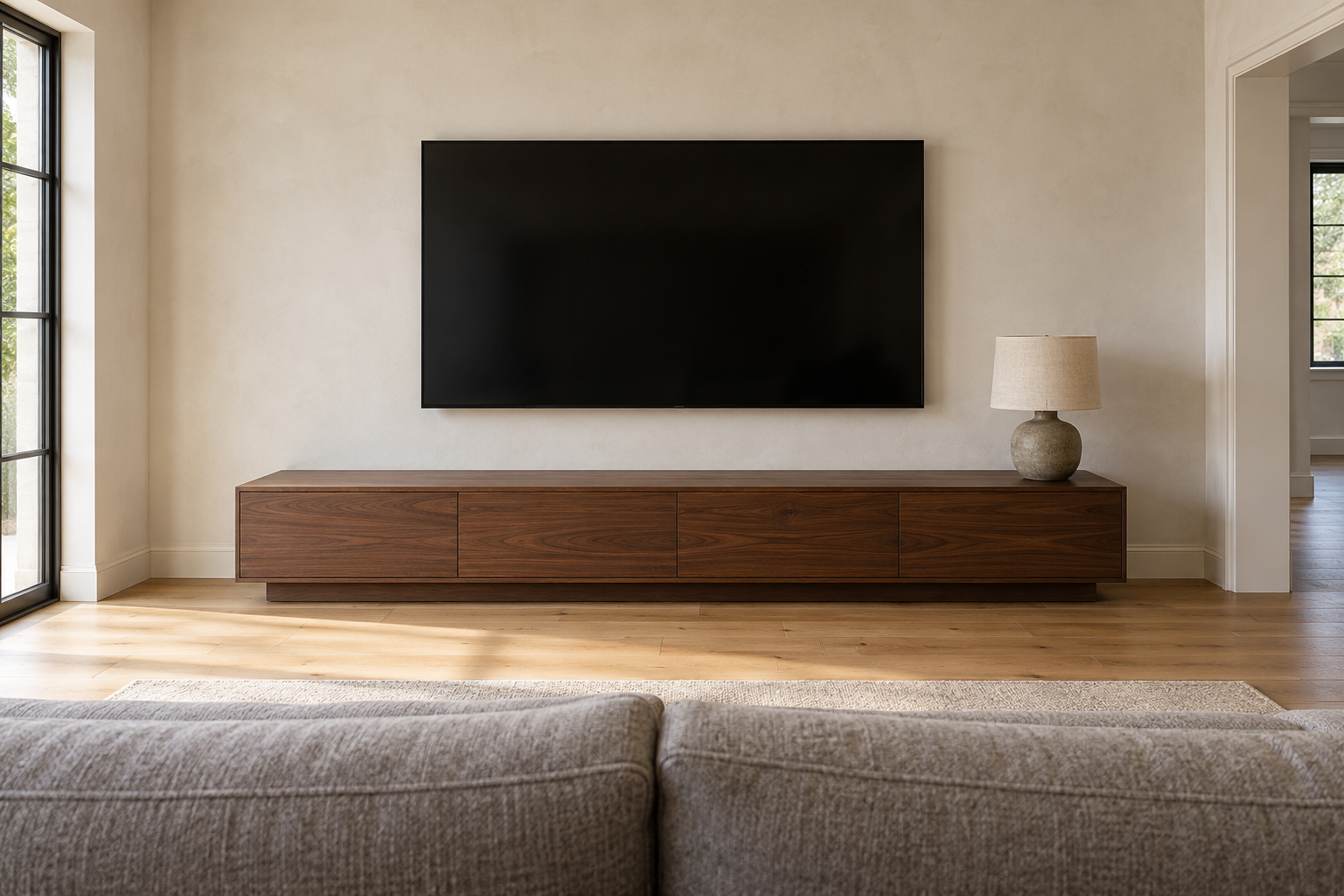 A dark television mounted on a pale plaster wall a few inches above a long low walnut console on a plinth, the console wider than the screen.