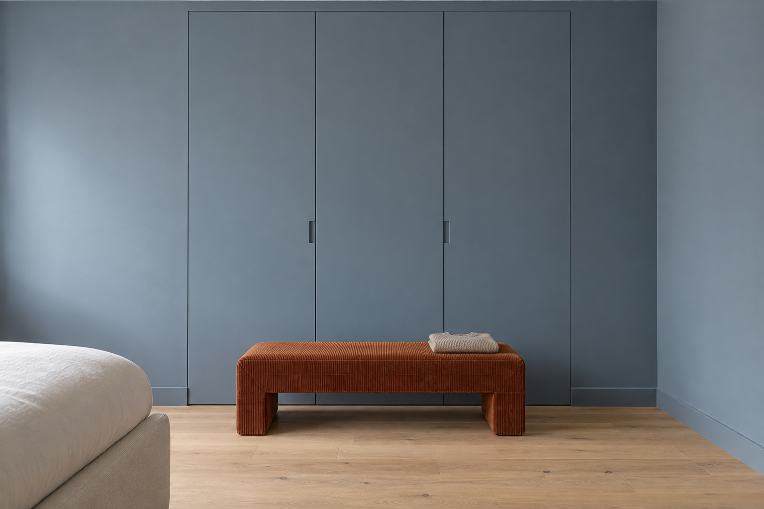 A full-height wardrobe painted the same blue-gray as the bedroom wall, with a rust bench in front.