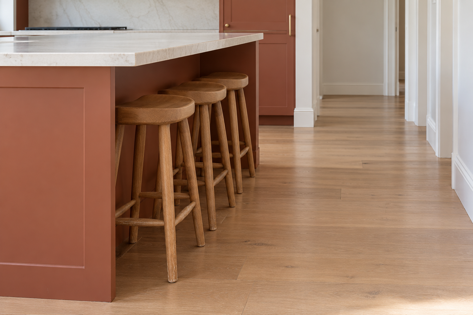 Three backless oak stools pushed fully under the overhang of a terracotta red island, with the aisle behind them clear.