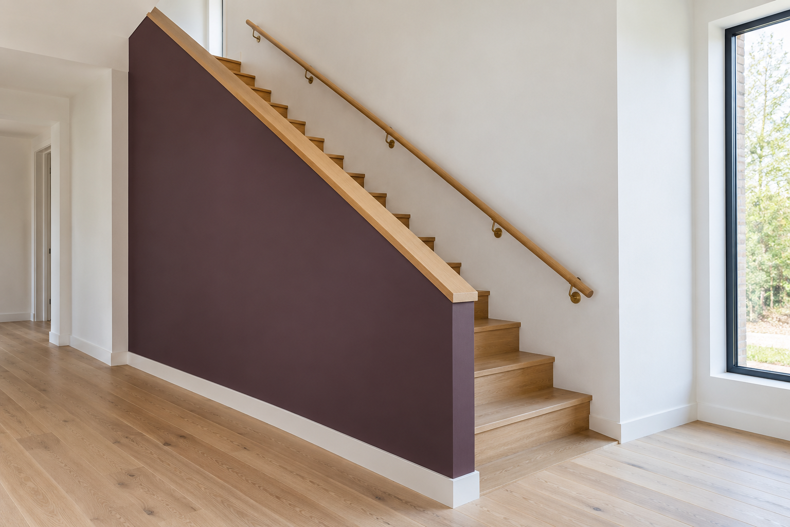 A staircase enclosed by a solid deep plum half wall with an oak cap, and a round oak handrail on brass brackets along the white wall on the other side of the stair.