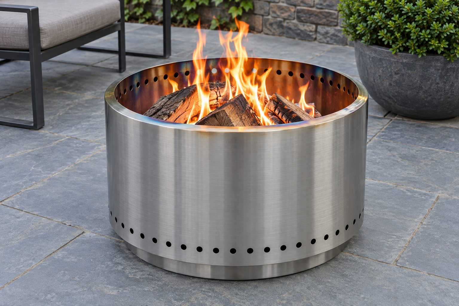 A brushed stainless steel cylinder fire pit on a bluestone patio, with a row of small holes visible inside its rim.