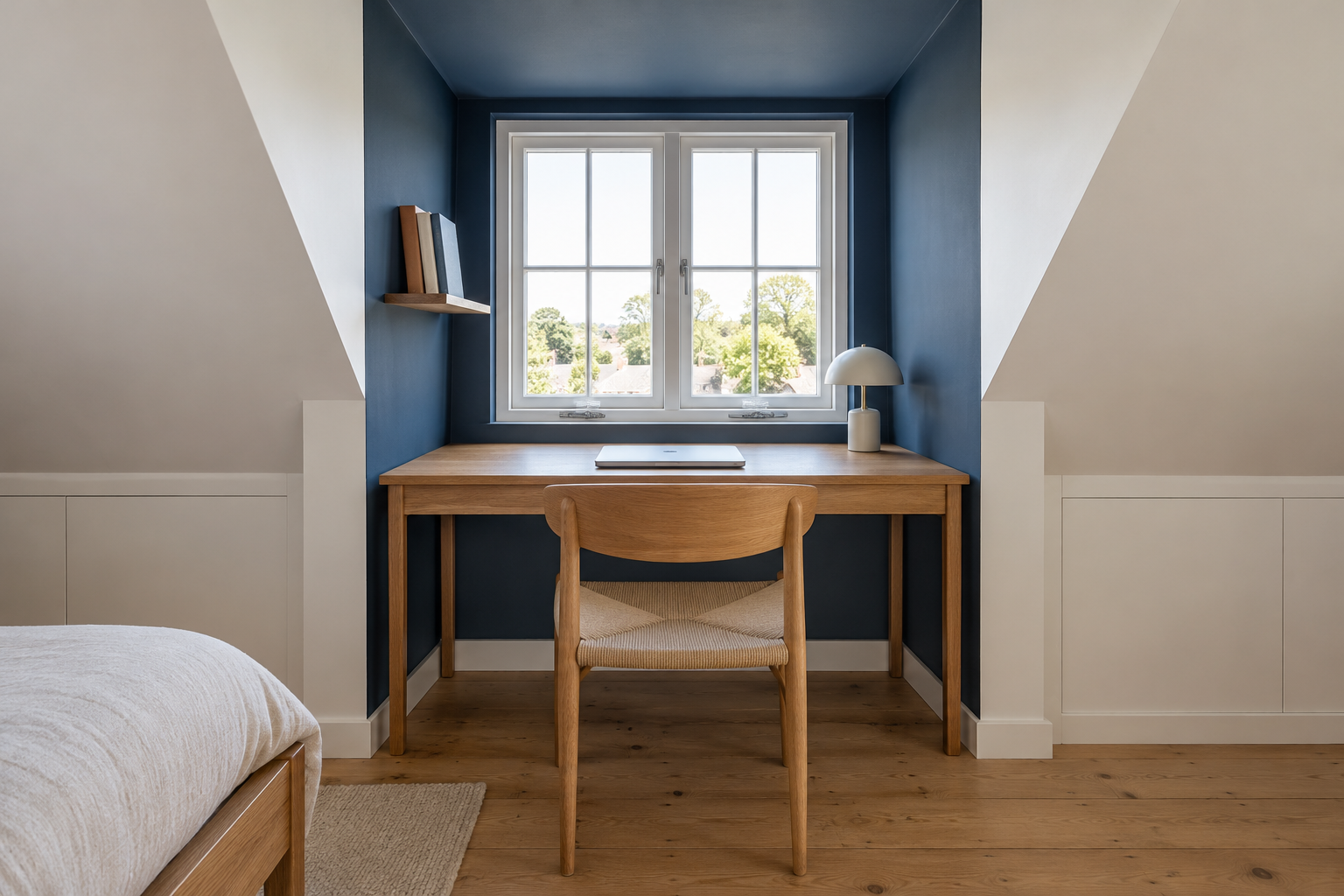 A small desk set into an attic dormer painted deep blue inside, facing the dormer window, with a chair pulled up under the dormer's flat ceiling.