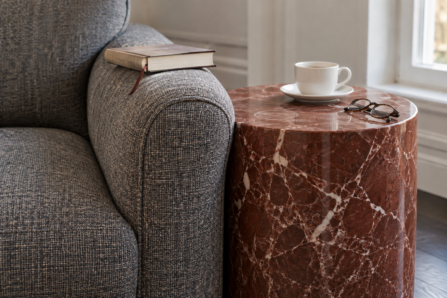 A small red marble drum table beside a gray tweed armchair, its top level with the chair arm, holding a cup and a pair of glasses.