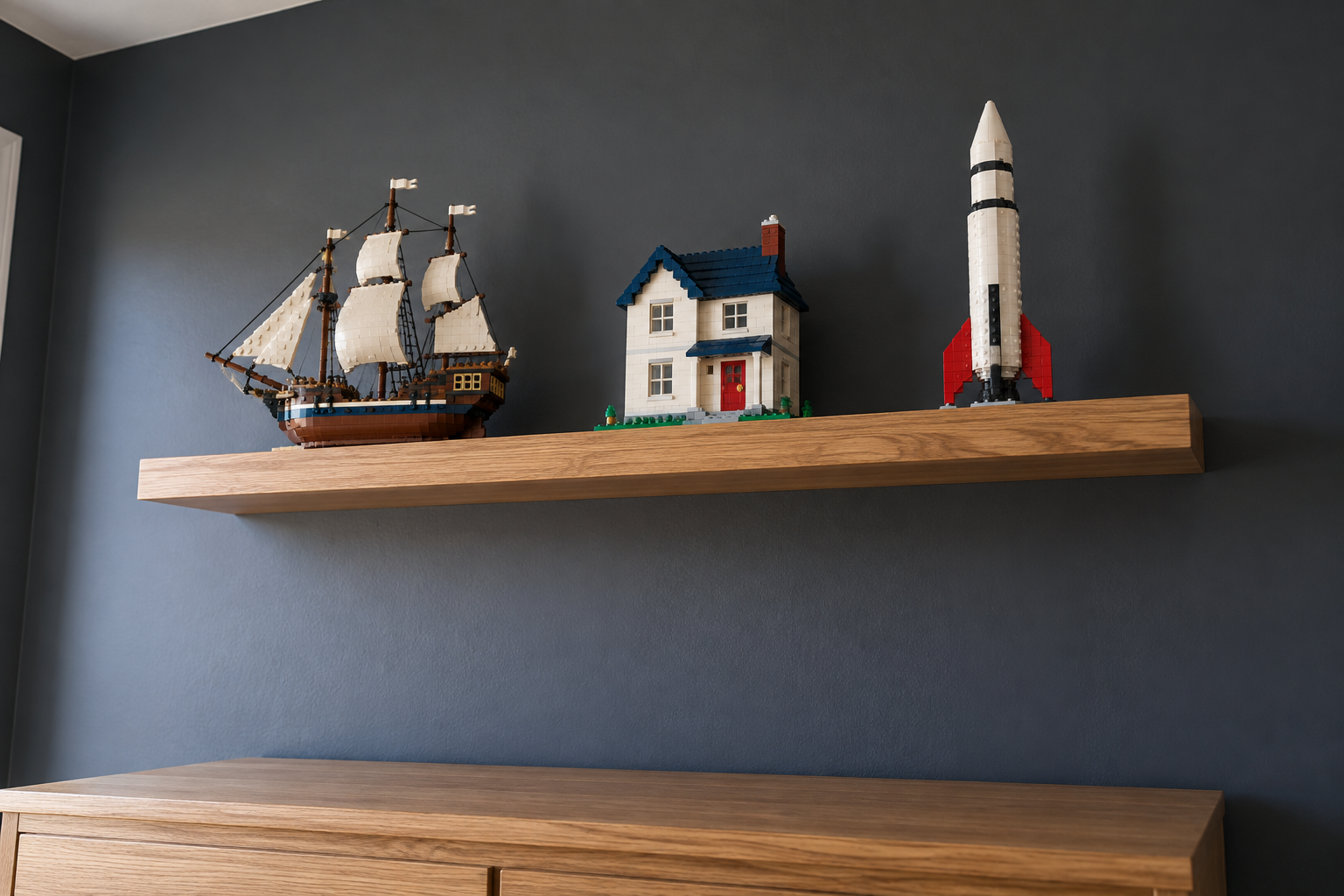 Three brick-built models, a ship, a house, and a rocket, on an oak shelf high on a charcoal wall.