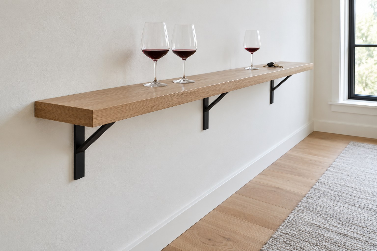 A narrow white oak ledge on slim black steel brackets running along a white wall beside a window at elbow height, holding three glasses and a set of keys.